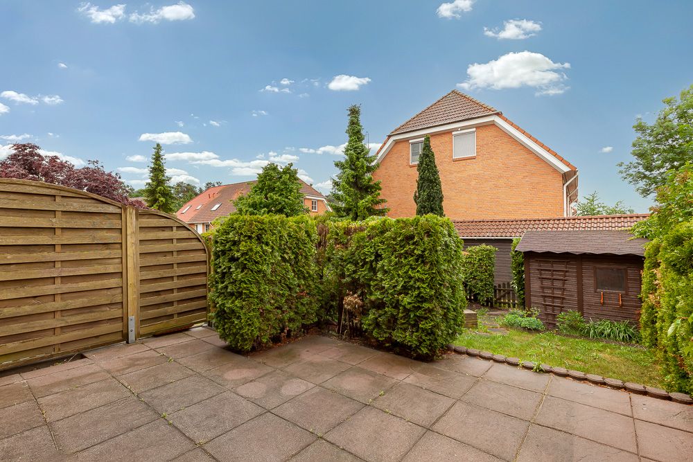 Gepflasterte Terrasse mit Hecke, Gartenhaus und Blick auf angrenzende Wohnhäuser.