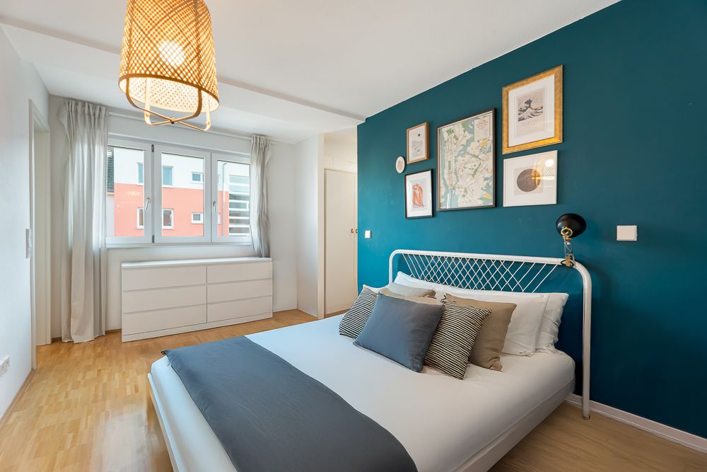 Helles Schlafzimmer mit Doppelbett, Kommode und Bildern an einer blauen Akzentwand.