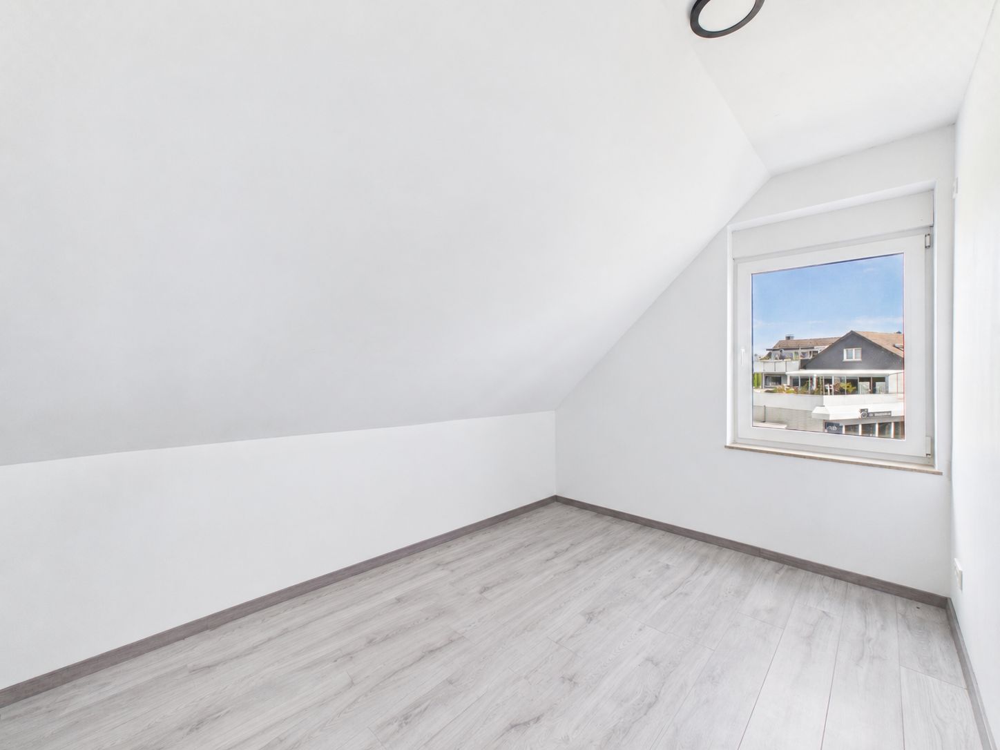 Helles, leeres Zimmer mit schräger Decke, Fenster und hellem Laminatboden.