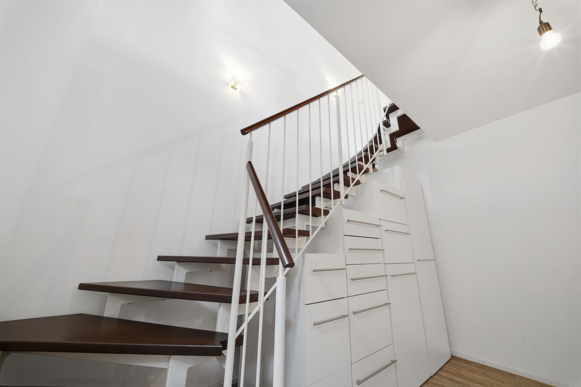Holztreppe mit weißem Geländer und Einbauschränken darunter in einem hellen Raum.