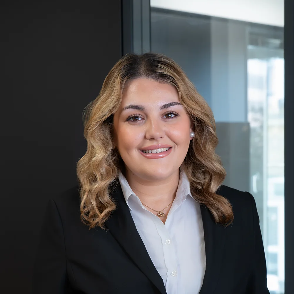 Frau mit lockigen blonden Haaren im schwarzen Blazer steht vor Glaswand.