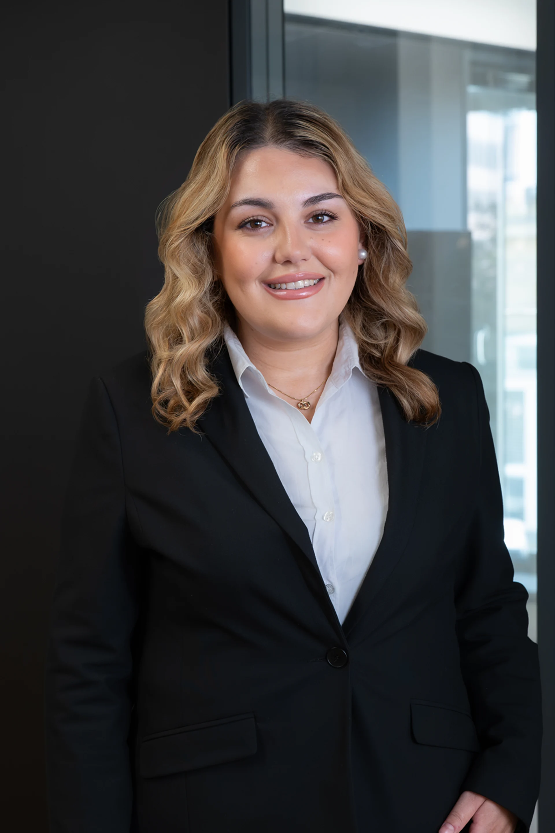 Frau mit lockigen blonden Haaren im schwarzen Blazer steht vor Glaswand.