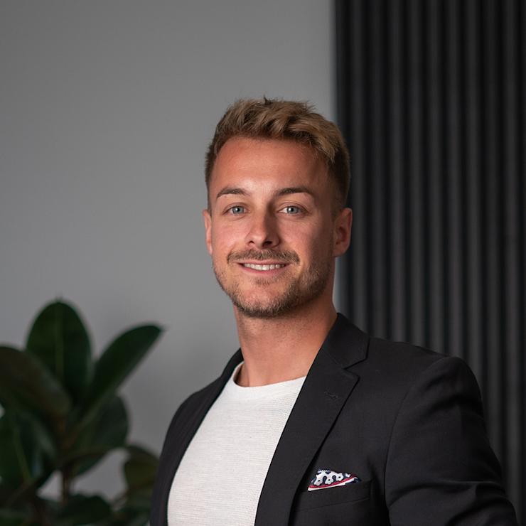 Ein junger Mann im Anzug steht vor einer Pflanze und einer Wand mit vertikalen Streifen.