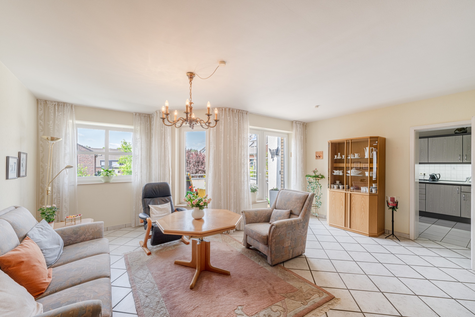 Helles Wohnzimmer mit Sofa, Sessel, Esstisch, Teppich, Vitrine und Zugang zur Küche.