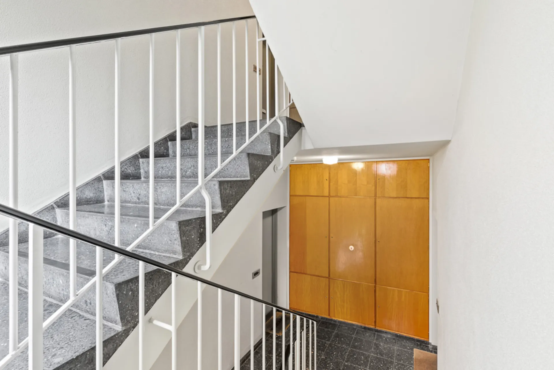 Treppenhaus mit grauer Steintreppe, weißem Geländer und Holztüren im Hintergrund.
