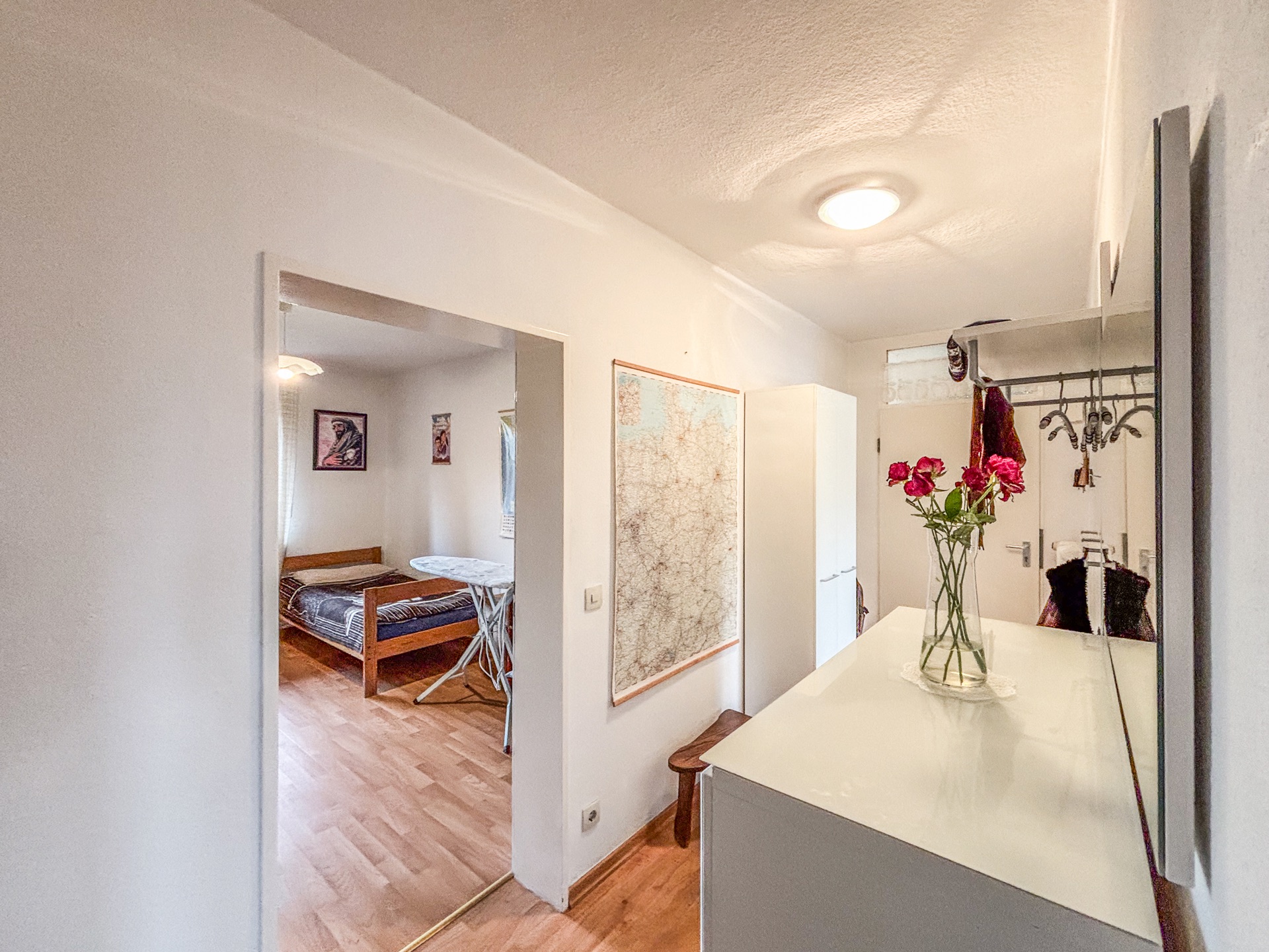 Flur mit Spiegel, Kommode, Blumen in Vase und Blick in Schlafzimmer mit Bett und Bügelbrett.