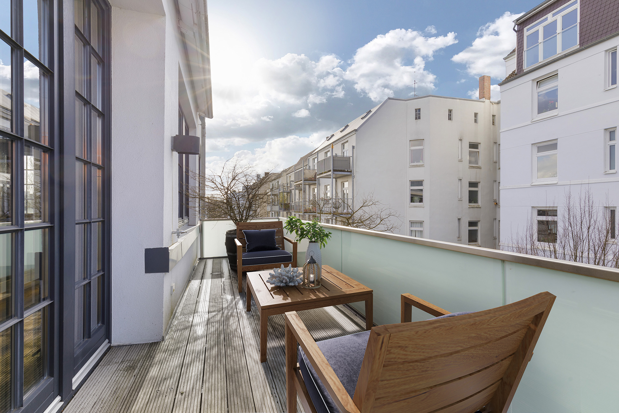 Möblierter Balkon mit zwei Sesseln, Tisch und Pflanzen, umgeben von Wohngebäuden bei Sonnenschein.