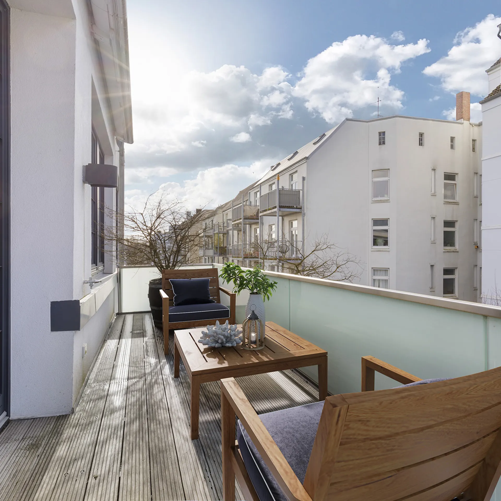 Möblierter Balkon mit zwei Sesseln, Tisch und Pflanzen, umgeben von Wohngebäuden bei Sonnenschein.