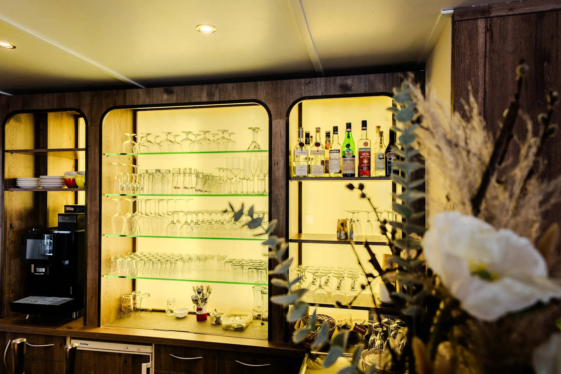 Hausbar mit Gläsern, Flaschen und Kaffeemaschine, davor ein Blumenstrauß.