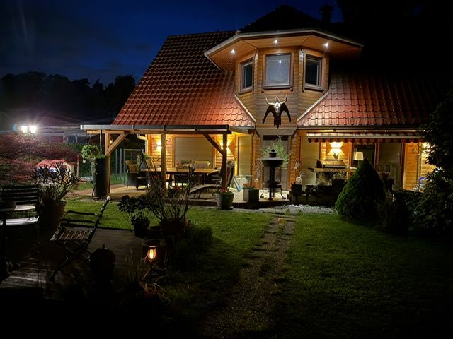 Beleuchtetes Holzhaus mit rotem Dach, Terrasse und Garten bei Nacht.