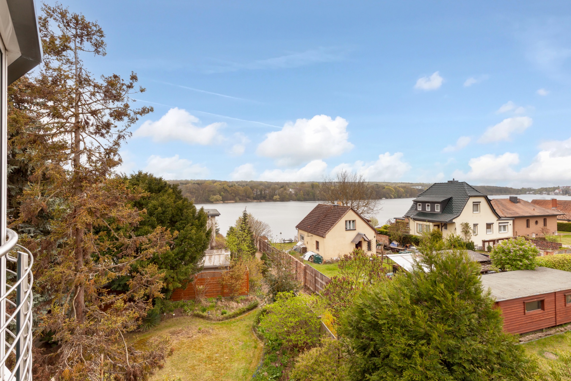 Blick auf einen Garten, mehrere Häuser und einen See im Hintergrund bei blauem Himmel.