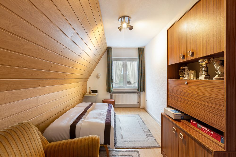 Schmales Schlafzimmer mit Einzelbett, Holzschrank, Sessel und Fenster mit Vorhängen.