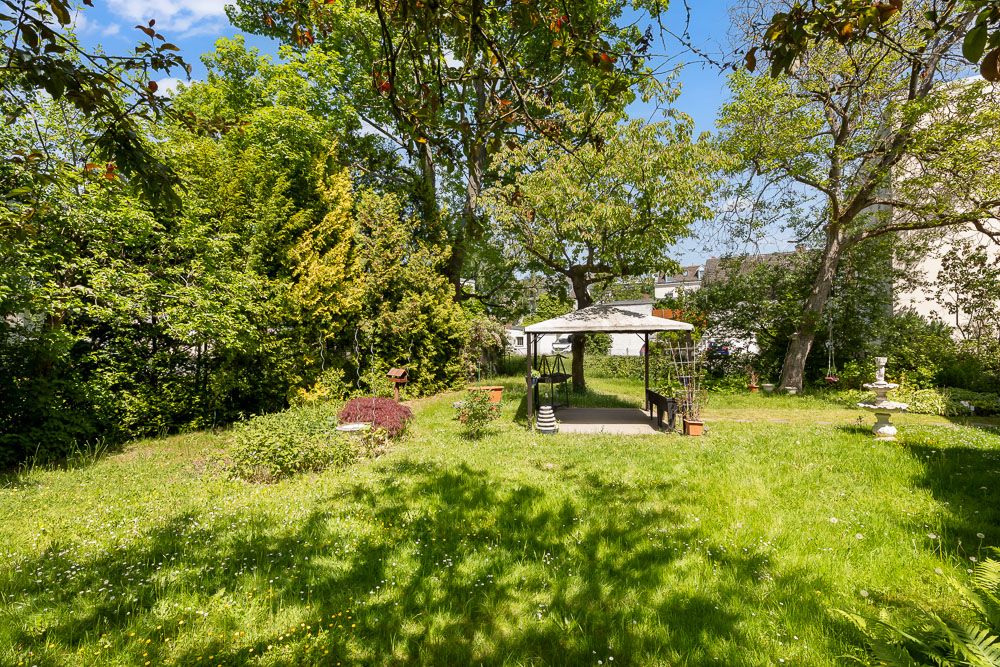 Grüner Garten mit Rasen, Bäumen und einem Pavillon mit Sitzbank in der Mitte.