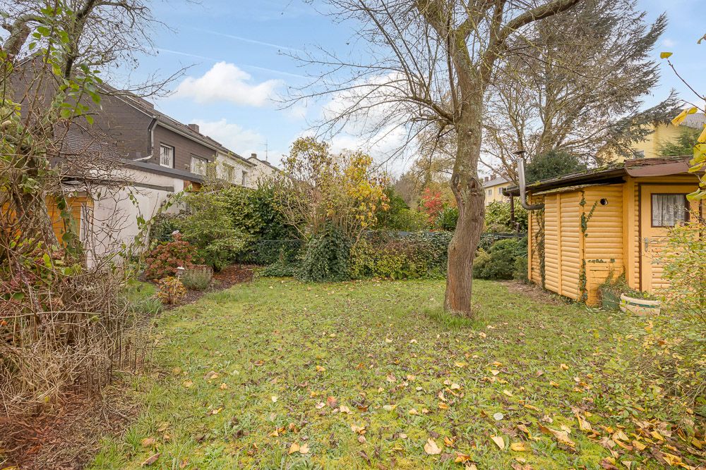 Garten mit Rasenfläche, Baum, Sträuchern und einem Holzhaus am rechten Rand.