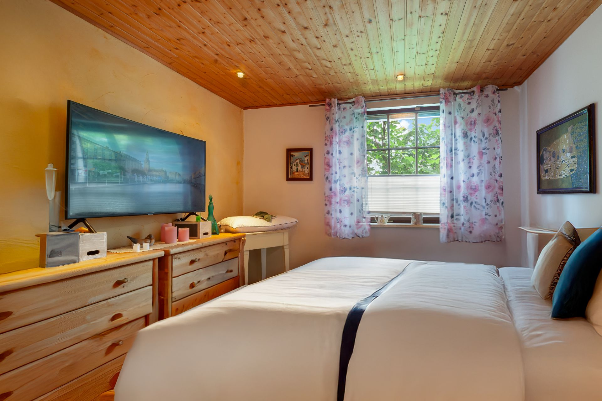 Schlafzimmer mit Doppelbett, Holzdecke, zwei Kommoden, TV und Fenster mit gemusterten Vorhängen.