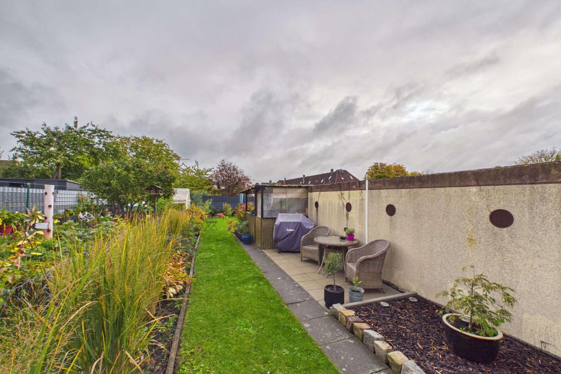 Langer Garten mit Rasen, Pflanzenbeeten, Sitzbereich und Gartenmöbeln an einer Mauer.