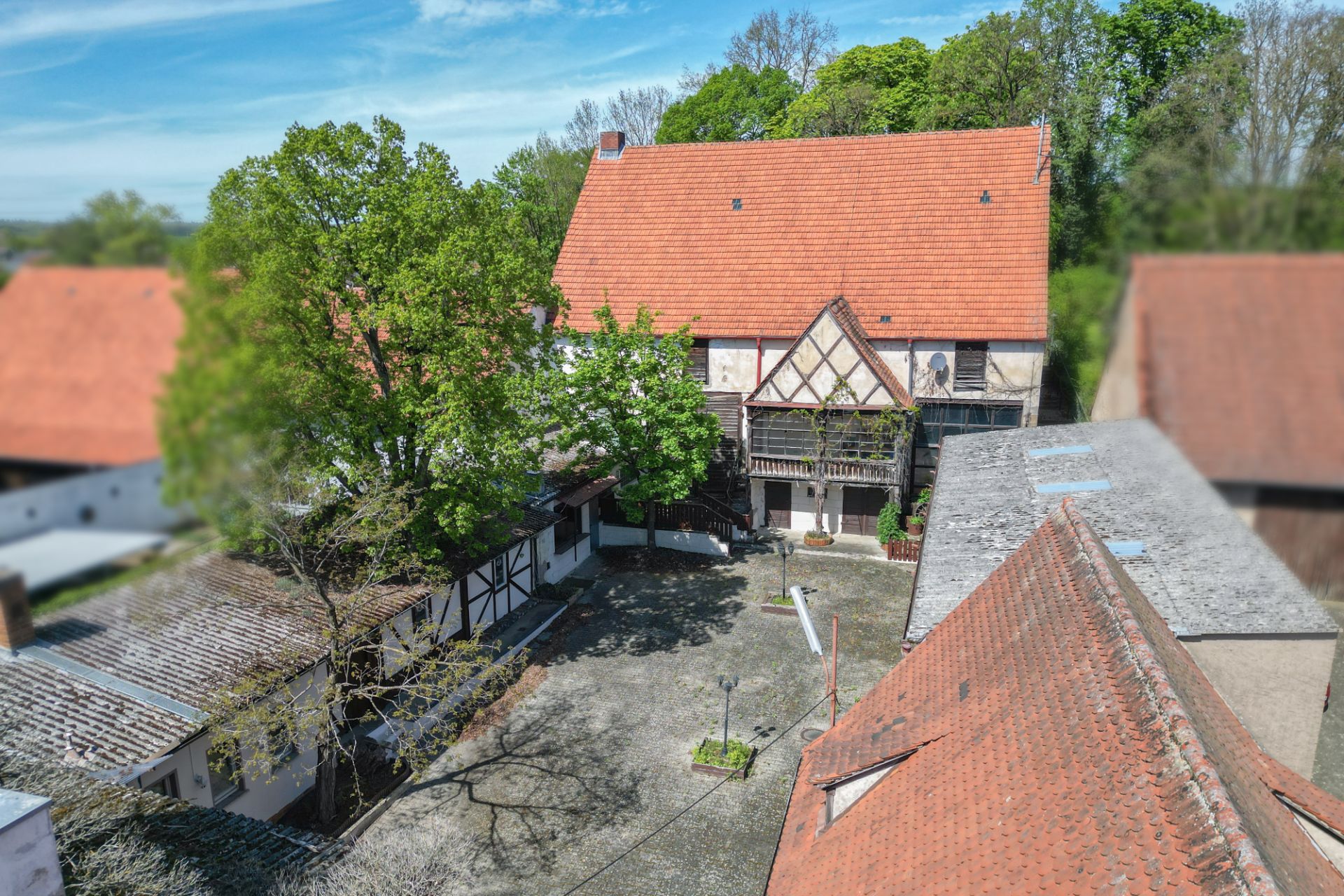 Innenhof mit Fachwerkhaus und roten Dächern, umgeben von Bäumen und Nebengebäuden.