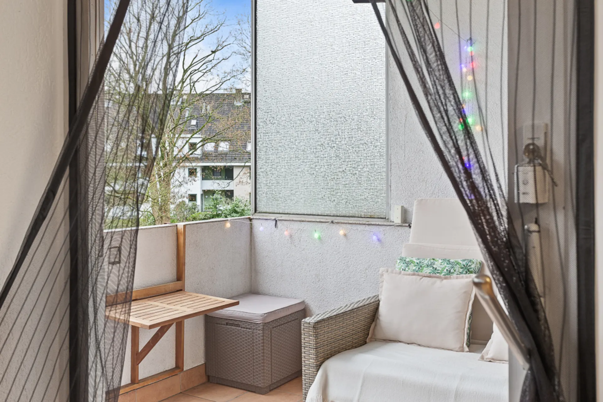 Kleiner Balkon mit Sitzbank, Kissen, Holztisch und bunter Lichterkette an der Wand.