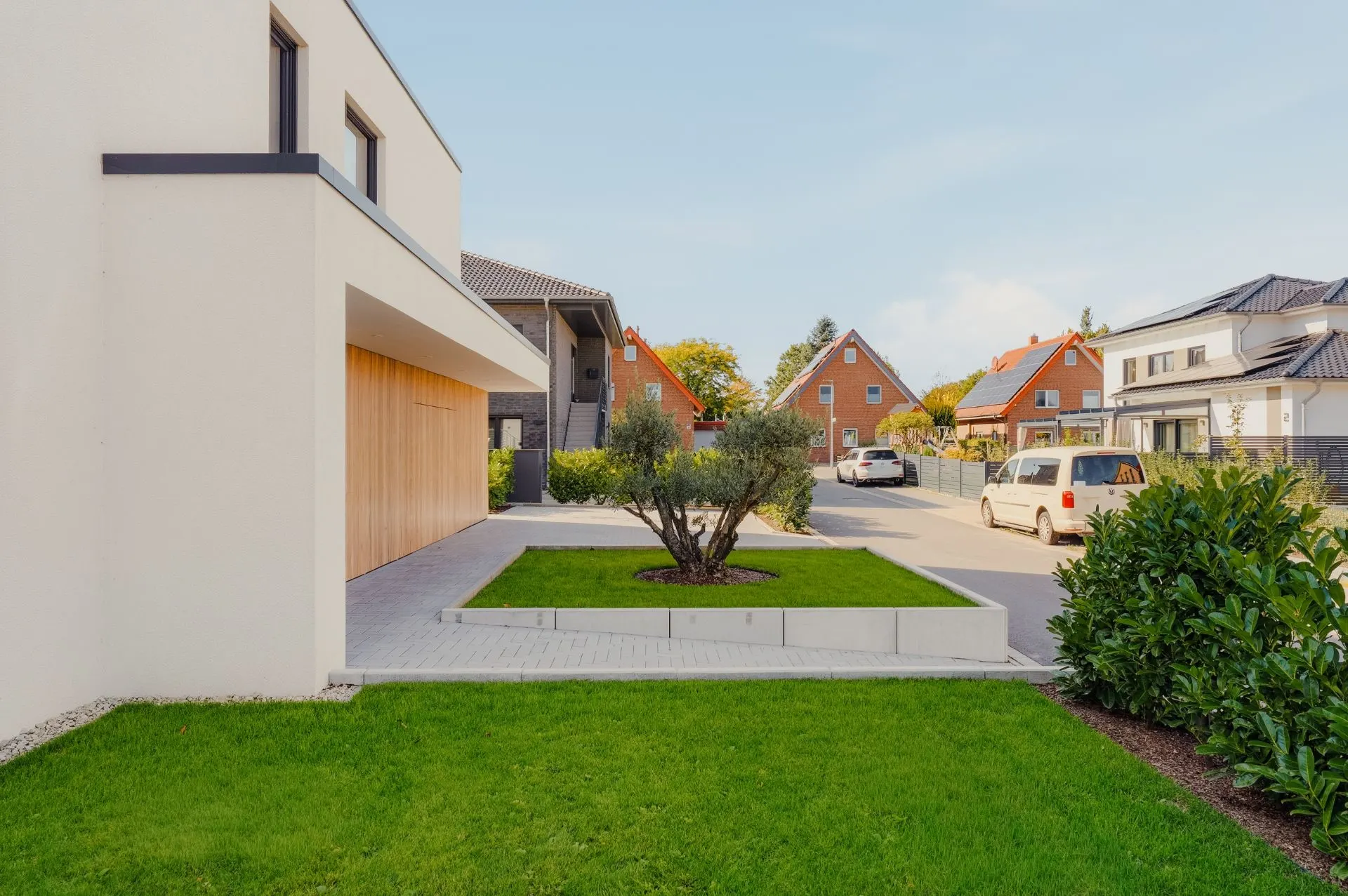 Moderner Wohnhausgarten mit Baum und Rasen, umliegende Nachbarhäuser und parkende Autos sichtbar.