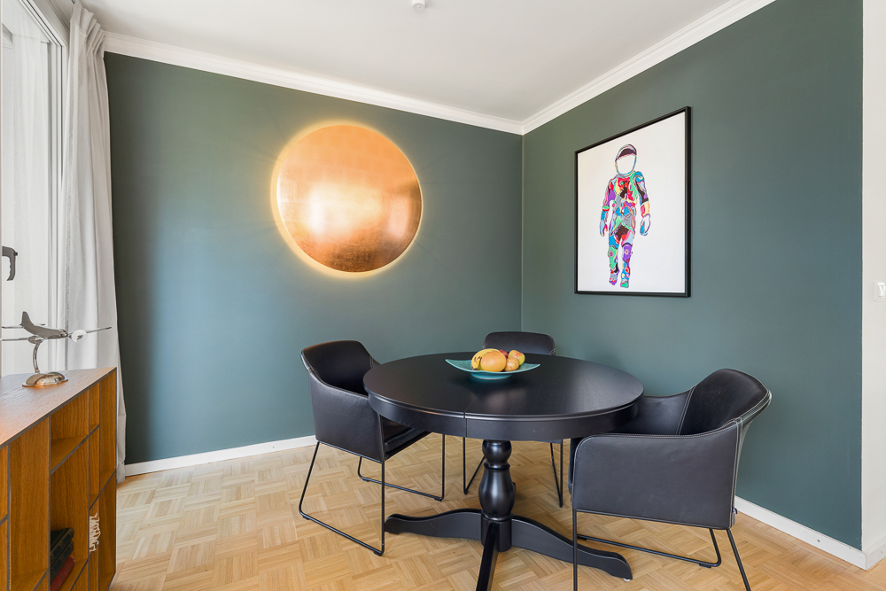 Essbereich mit rundem schwarzen Tisch, vier Stühlen, Kunstwerk und goldener Wandlampe.