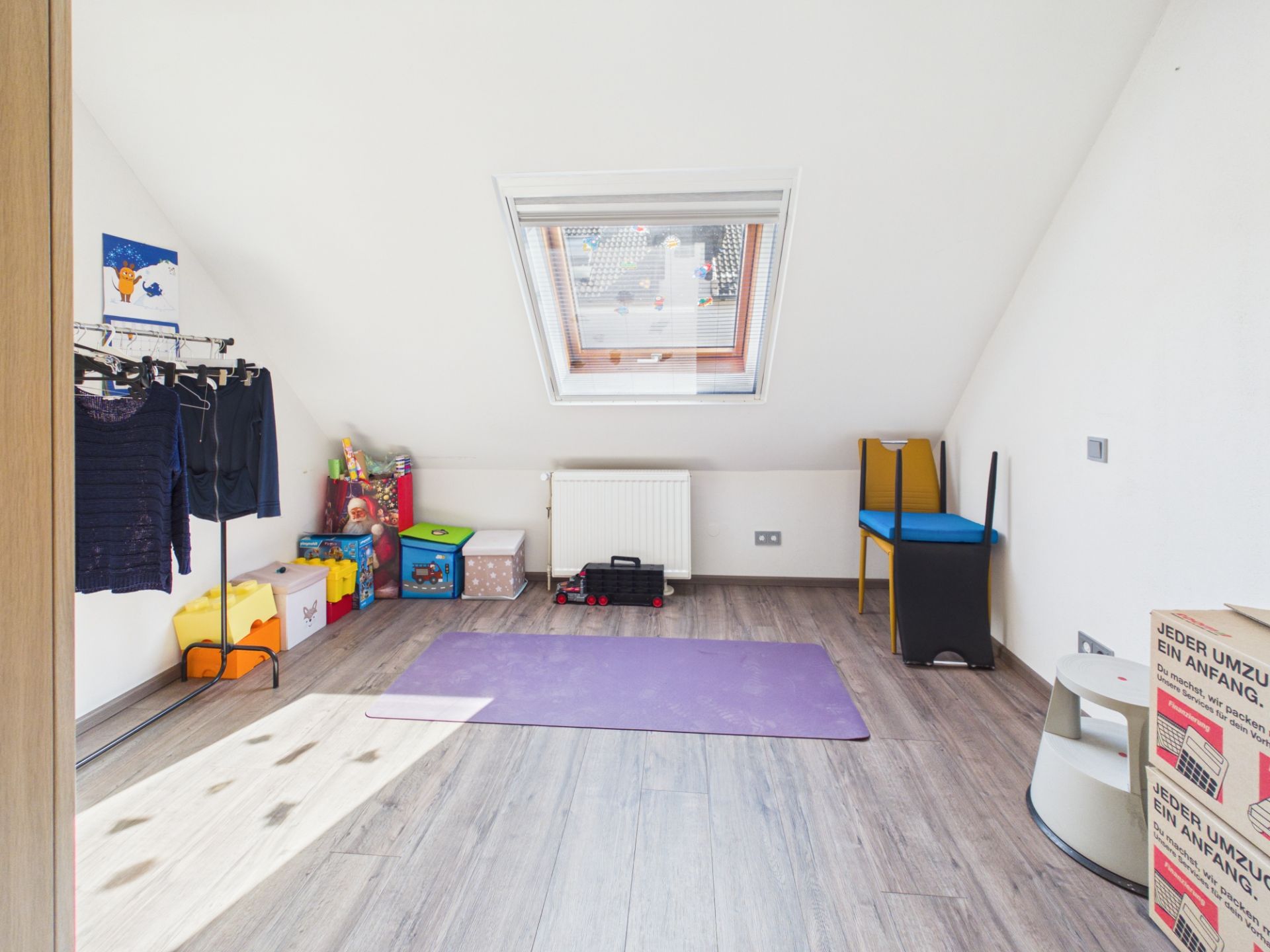 Helles Zimmer mit Dachfenster, Spielzeugkisten, Kleiderstange und Umzugskartons.