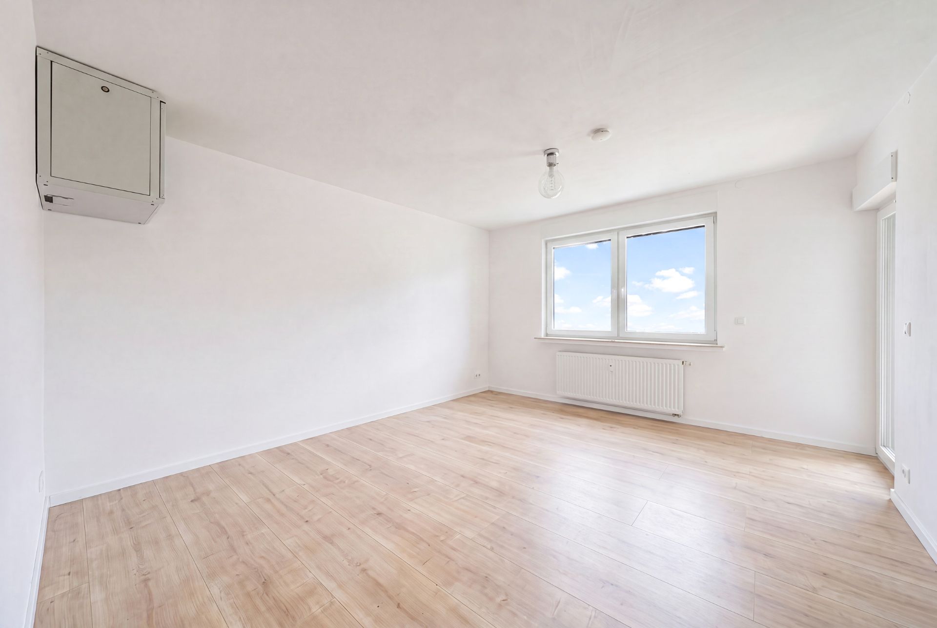 Leerer, heller Raum mit Holzfußboden und großem Fenster, weiße Wände.