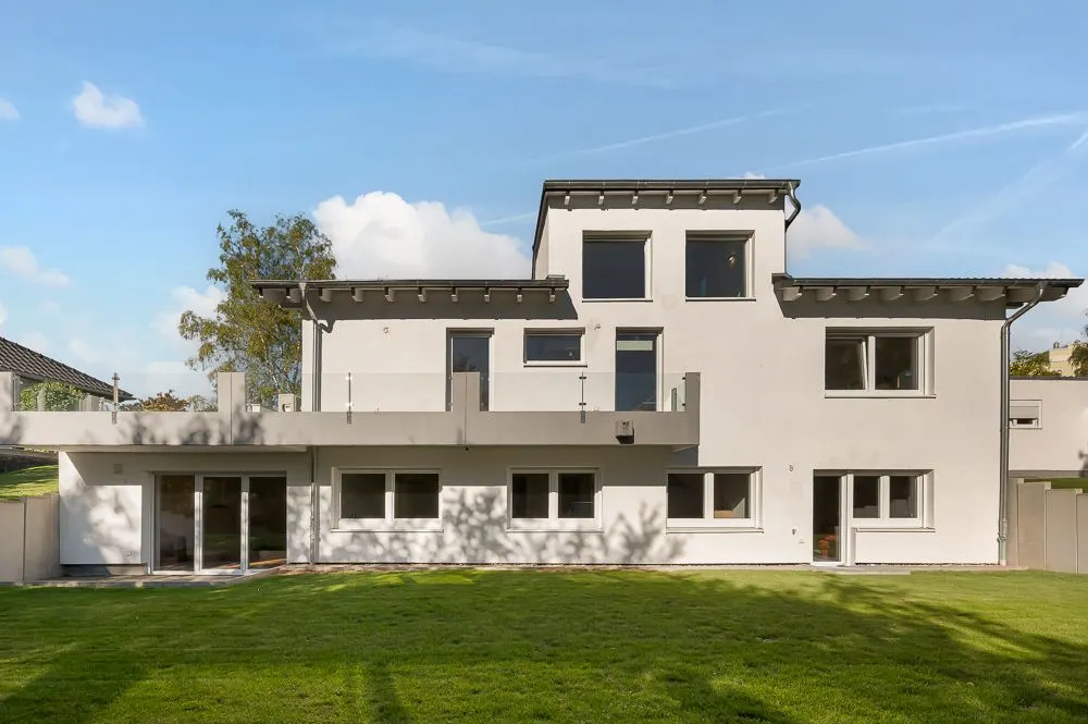 Moderne weiße Hausfassade mit vielen Fenstern und Türen, davor eine grüne Rasenfläche.