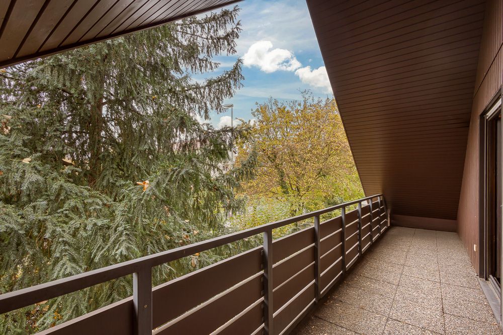 Überdachter Balkon mit braunem Geländer und Blick auf grüne Bäume.
