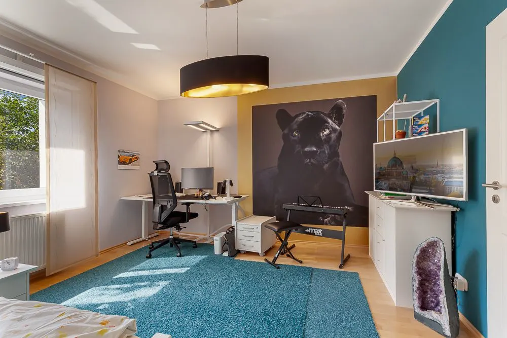 Helles Zimmer mit Schreibtisch, Stuhl, großem Panther-Bild, Teppich und Keyboard.