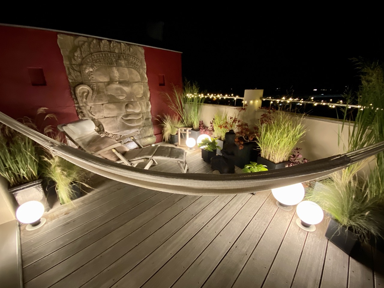 Beleuchtete Terrasse mit Hängematte, Pflanzen, Liegestuhl und großer Reliefskulptur an der Wand.
