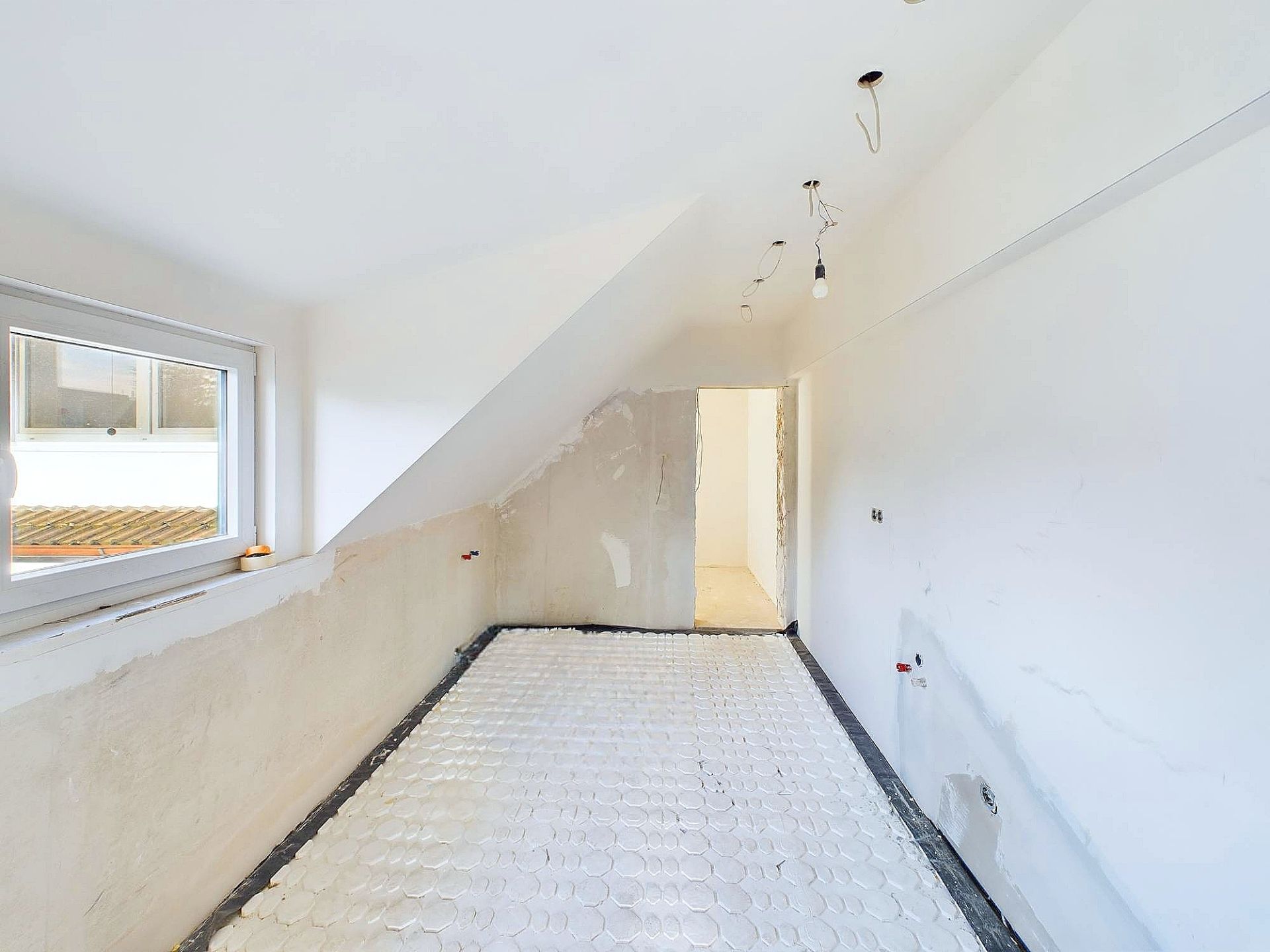 Leerer, renovierungsbedürftiger Raum mit Dachschräge, Fenster und offenliegenden Leitungen.