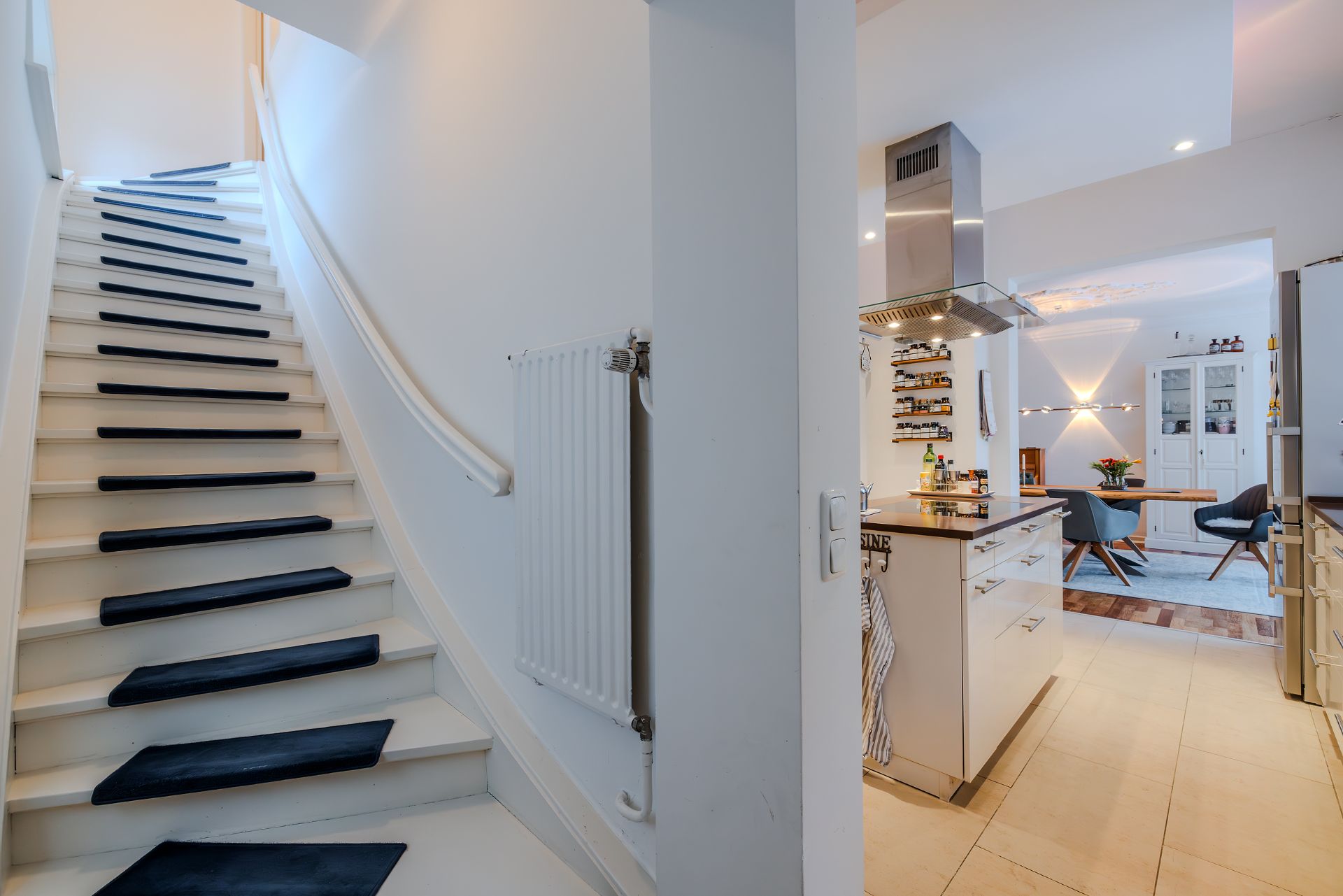 Weiße Treppe mit schwarzen Stufen, daneben Küche und Essbereich mit Tisch und Stühlen.