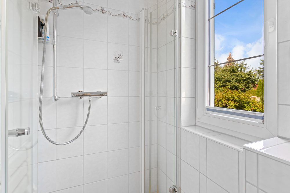 Weiße geflieste Dusche mit Glastür und Fenster mit Blick ins Grüne.