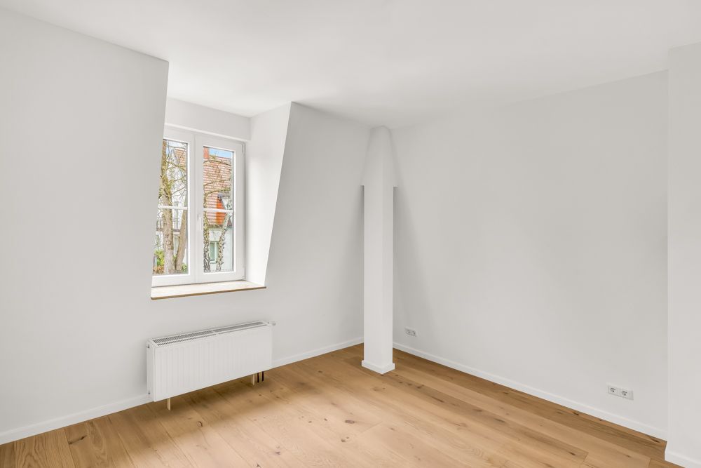 Helles, leeres Zimmer mit weiß gestrichenen Wänden, Holzboden und Fenster mit Heizkörper darunter.