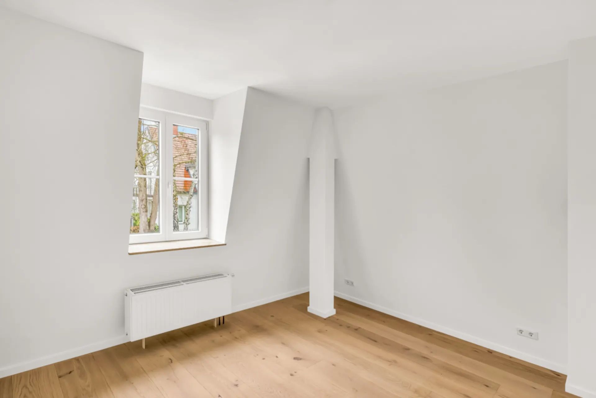 Helles, leeres Zimmer mit weiß gestrichenen Wänden, Holzboden und Fenster mit Heizkörper darunter.