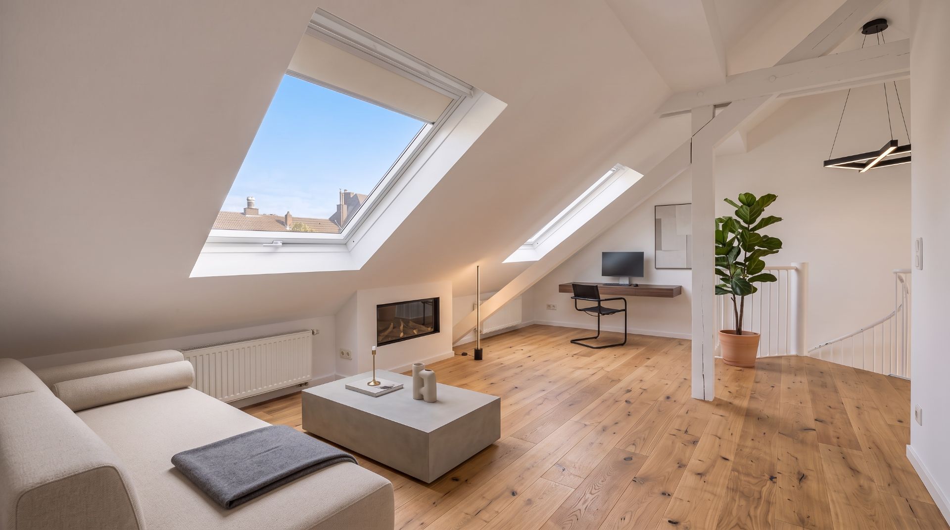 Helles Wohnzimmer mit Dachfenstern, Holzfußboden, Sofa, Couchtisch, Schreibtisch und großer Zimmerpflanze.