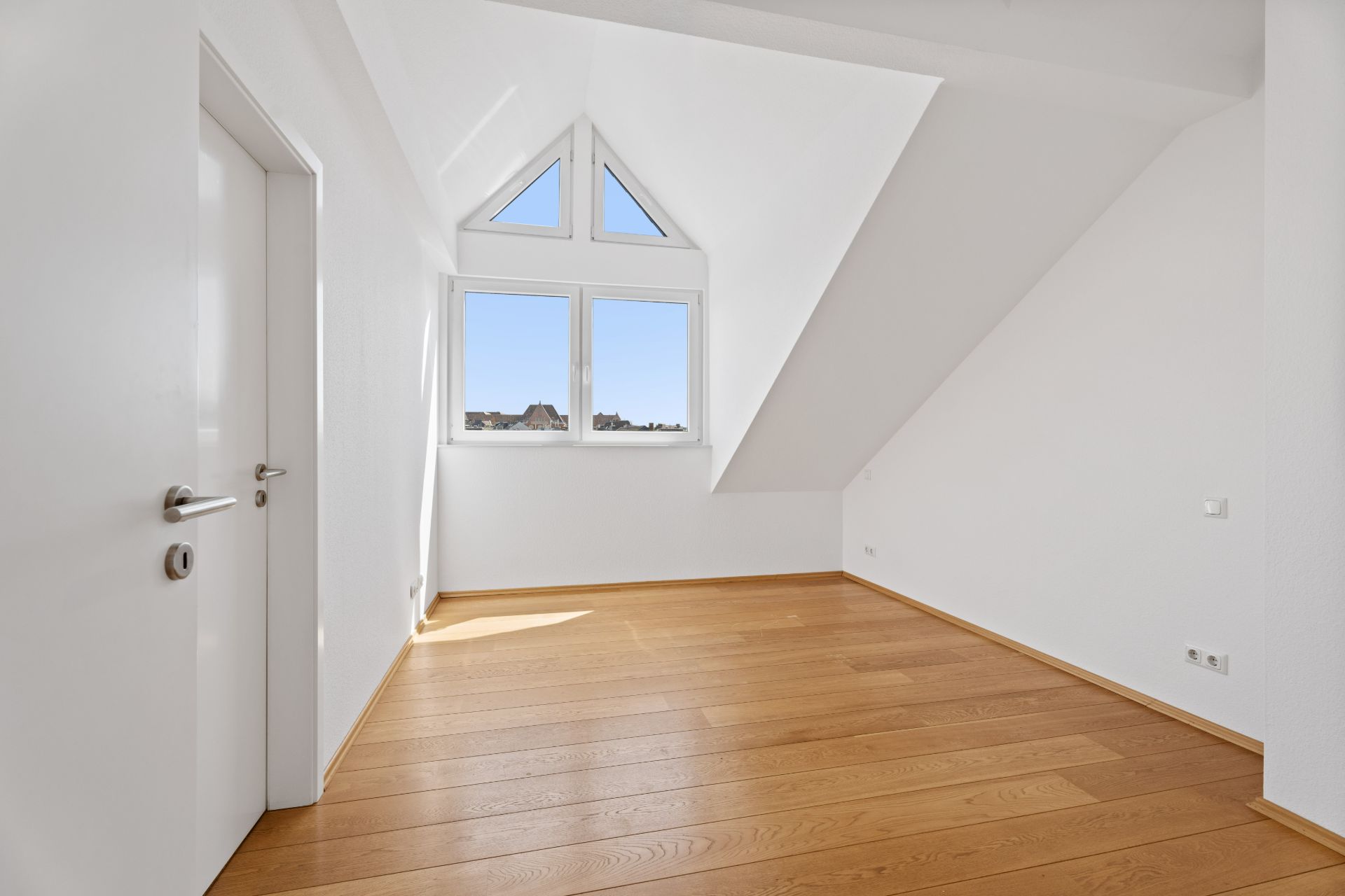 Leeres Zimmer mit weißer Wand, Holzboden und großen Fenstern unter dem Dach.