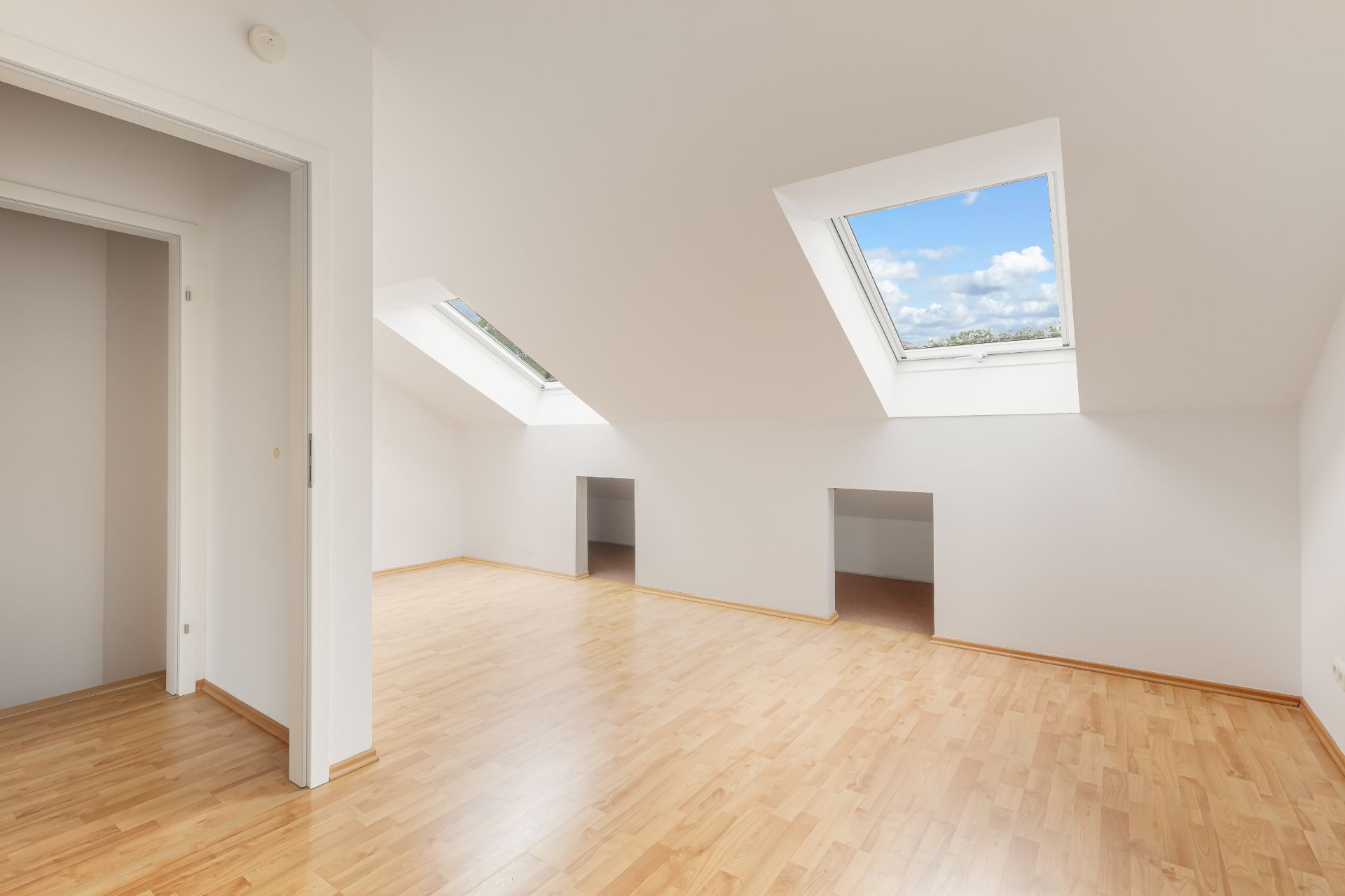 Heller Dachraum mit zwei Dachfenstern, Parkettboden und Zugang zu zwei kleinen Abstellräumen.