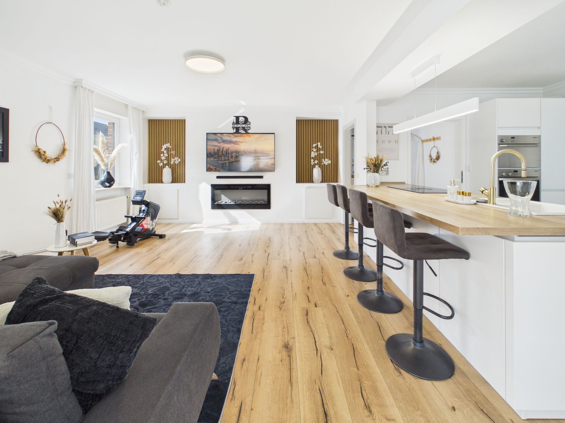 Helles Wohnzimmer mit Sofa, TV, Kamin, Kücheninsel und drei Barhockern auf Holzfußboden.