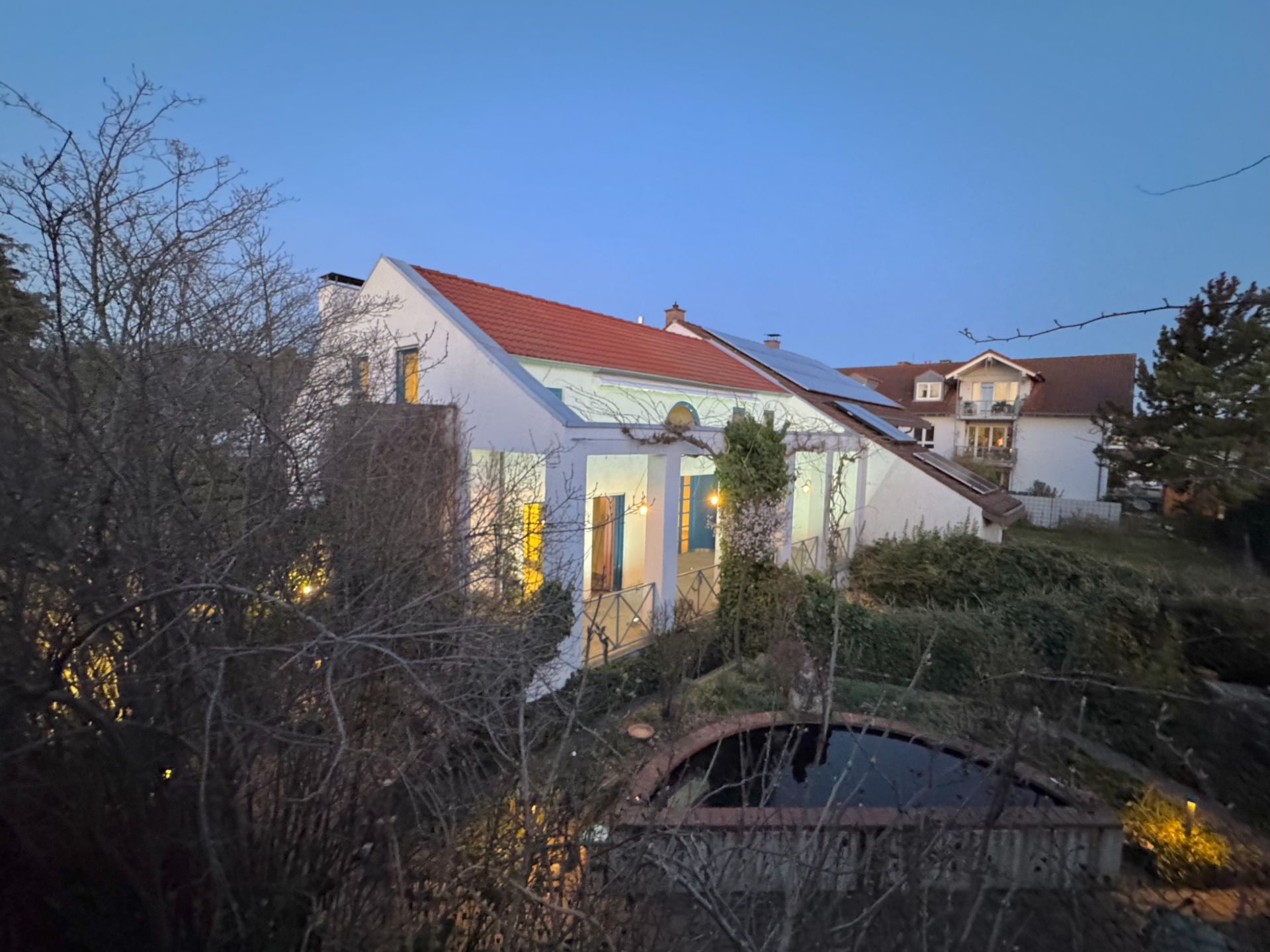 Weißes Haus mit roten Dachziegeln, Garten und kleinem Teich bei Dämmerung.