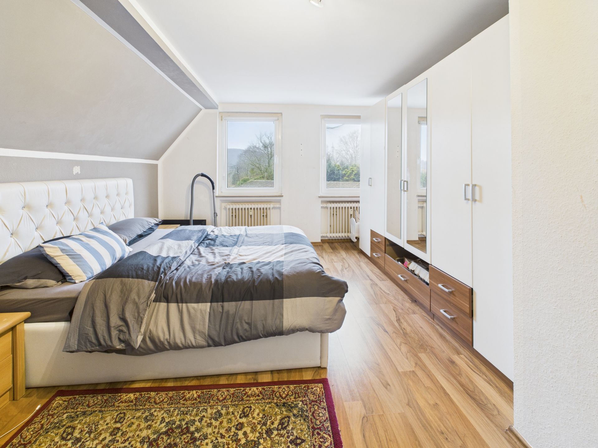 Schlafzimmer mit Doppelbett, Kleiderschrank, Holzboden und zwei Fenstern.