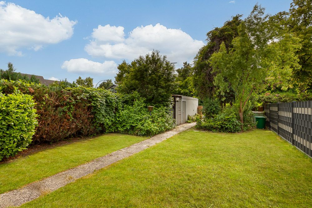 Gepflegter Garten mit Rasen, Gehweg und kleinen Gartenhaus, umgeben von Büschen und Bäumen.