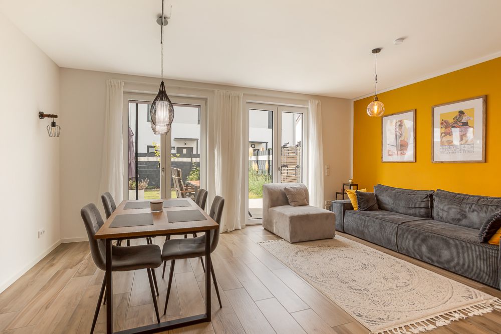 Helles Wohnzimmer mit gelber Wand, grauem Sofa, Esstisch und Zugang zum Garten.