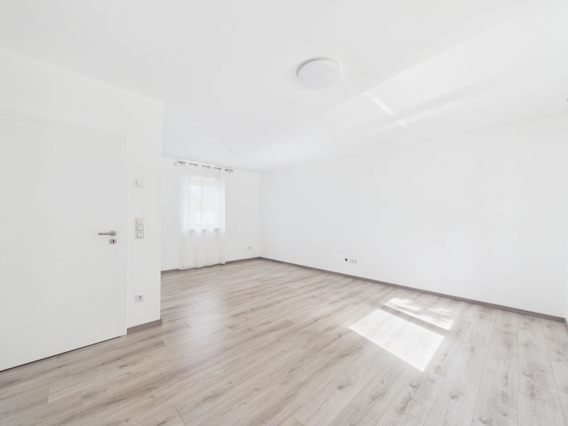 Helles, leeres Zimmer mit weißer Wand, Holzboden und einem Fenster mit Gardine.