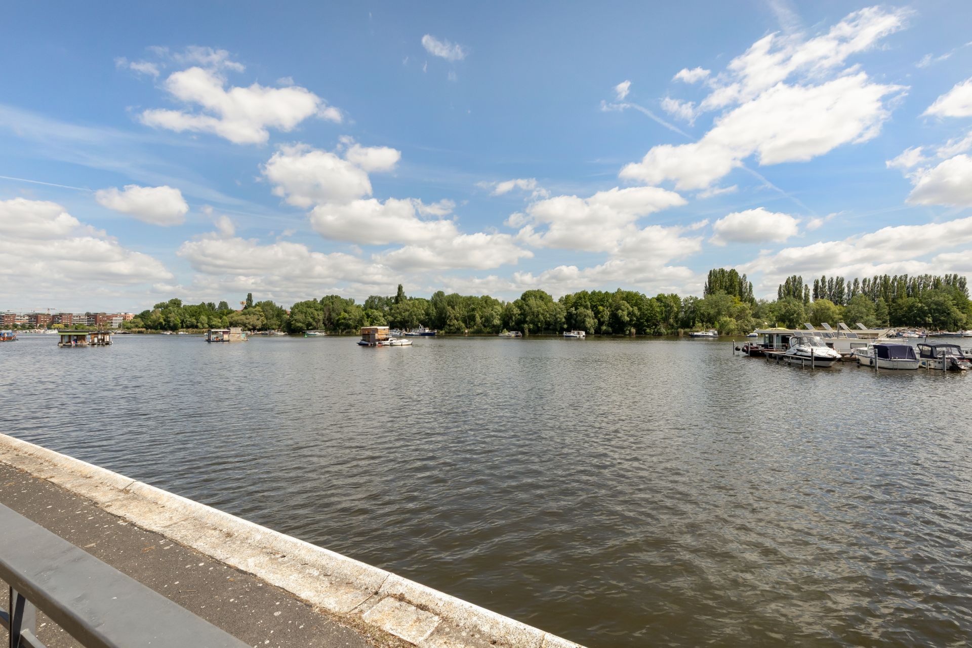 Blick von einer Uferpromenade auf einen Fluss mit Booten und Bäumen im Hintergrund.