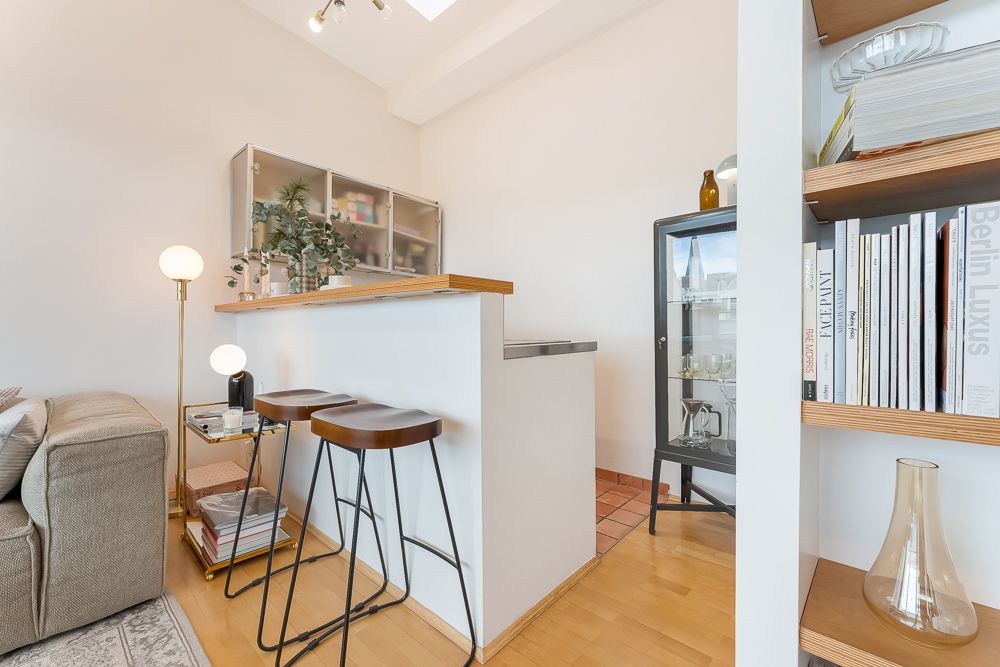 Wohnbereich mit Theke, zwei Hockern, Stehlampen, Sofa, Bücherregal und Vitrinenschrank.