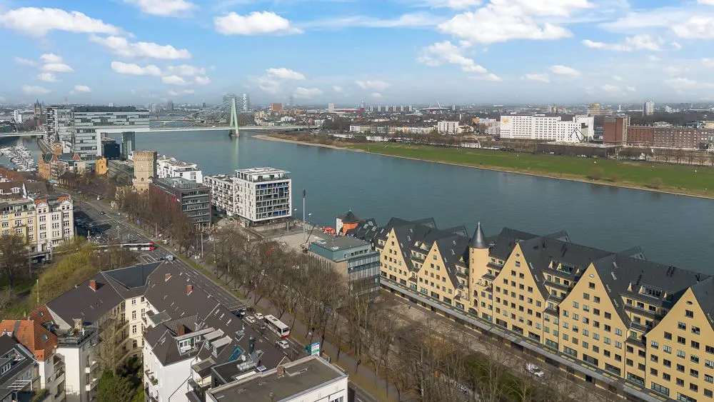 Blick auf Wohngebäude am Flussufer mit moderner Brücke und Park im Hintergrund.