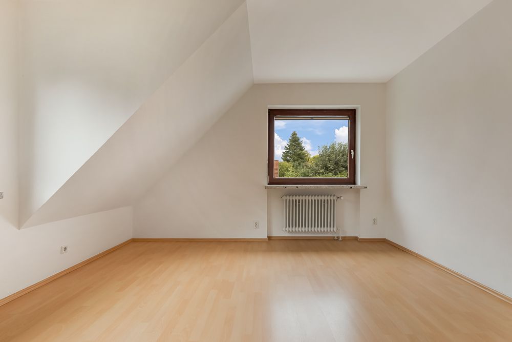 Leeres Zimmer mit hellem Laminatboden, weißer Wand und Fenster mit Blick ins Grüne.