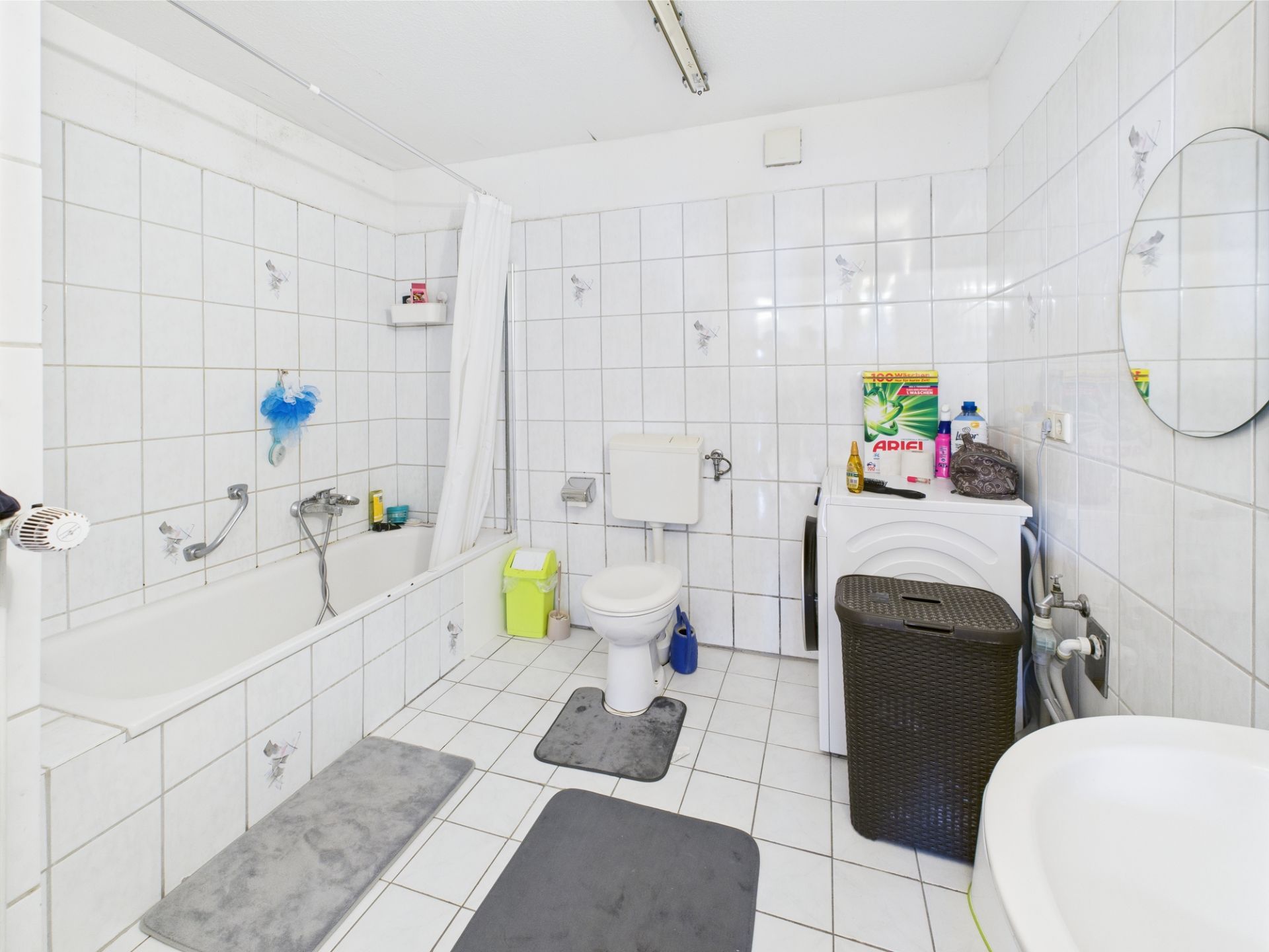 Helles Badezimmer mit Badewanne, Toilette, Waschmaschine und rundem Spiegel.