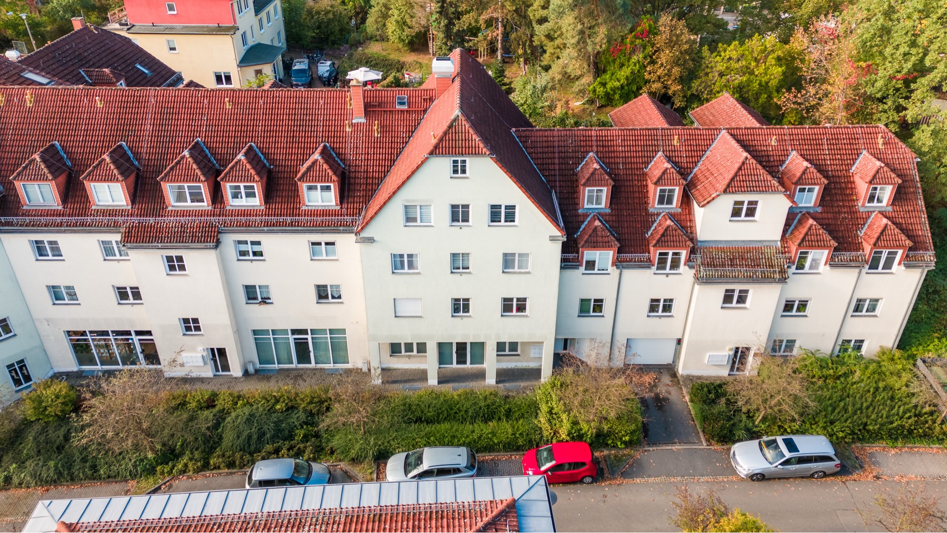 Mehrfamilienhaus mit rotem Dach, mehreren Fenstern und geparkten Autos davor.